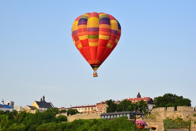 Een grote veelkleurige heteluchtballon vliegt laag over oude gebouwen en een dichtbeboste vallei. Van dichtbij gefotografeerd