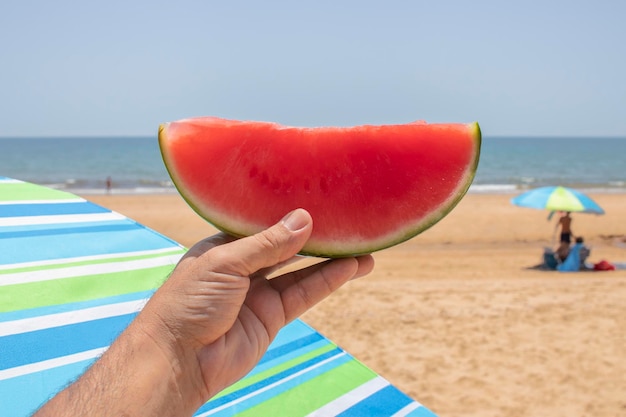 Een hand die een stuk watermeloen vasthoudt, op het strand. Concept van de natuurlijke manier om jezelf op te frissen..