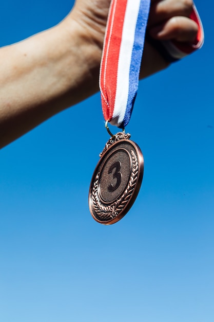 Een hand houdt een bronzen medaille op de derde plaats omhoog, met de lucht op de achtergrond. Overwinning concept