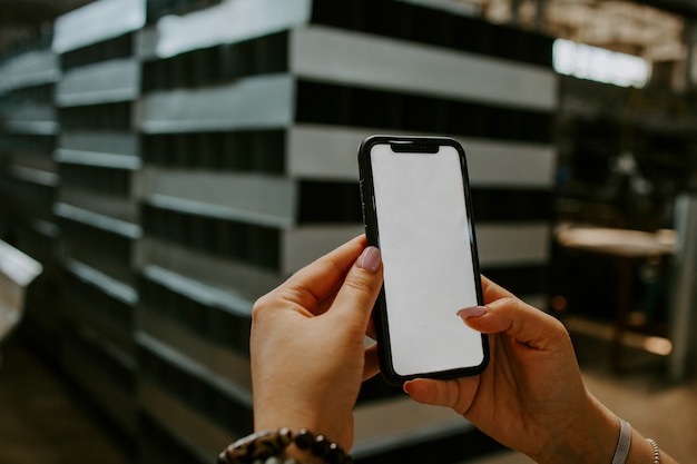 Een hand houdt een witte mobiele telefoon vast met een leeg wit scherm op de achtergrond van de fabriek