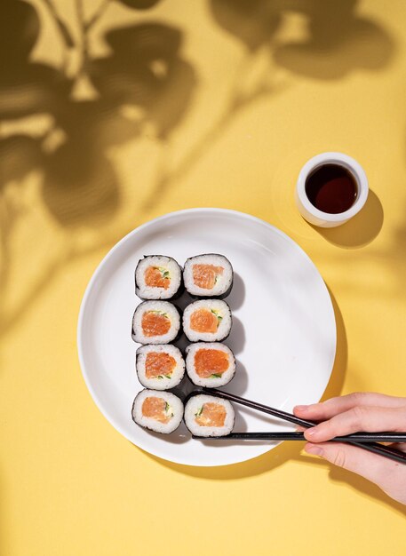 Een hand houdt eetstokjes vast met Japanse sushi-broodjes op een wit bord en sojasaus op een gele achtergrond met een harde schaduwbrunch Bovenaanzicht