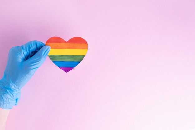 Een hand in een beschermende blauwe handschoen houdt een hart van regenboogpapier vast op een roze achtergrond Veilig Valentijnsdagconcept