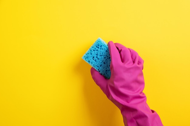 Een hand in een roze rubberen handschoen houdt een blauwe wasspons op een gele achtergrond