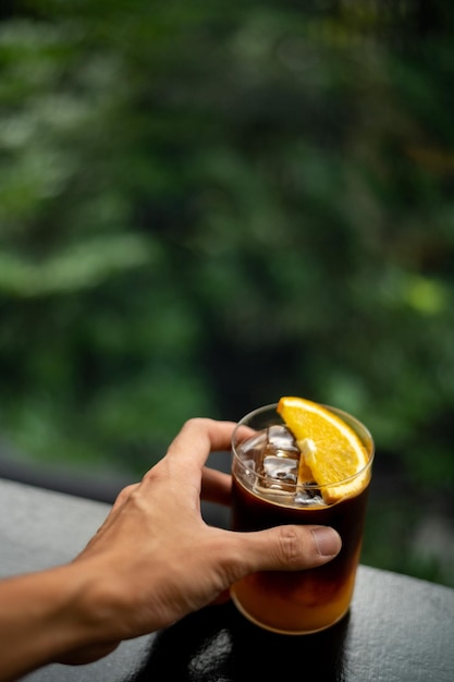 Een hand met een glas donkere cocktail met stukjes sinaasappel op de rand.