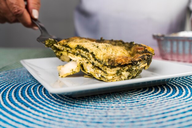 Een hand van een volwassen vrouw die een portie vers gemaakte pesto-lasagne op een bord serveert