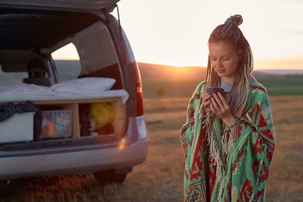 Een hippiemeisje in een lichte deken en met dreadlocks die koffie drinkt in het veld bij haar auto de ...