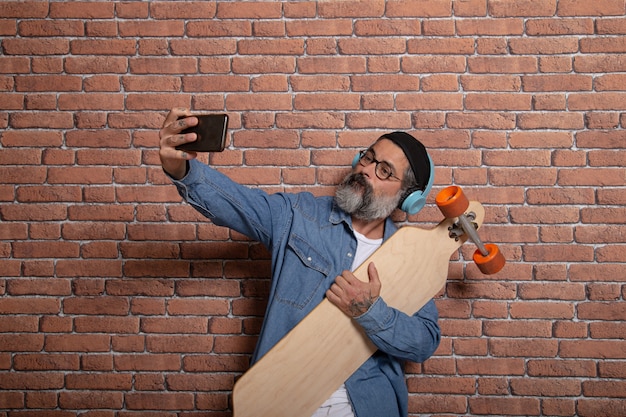 Een hipster maakt een selfie op zijn smartphone met het longboard op een bakstenen muur.