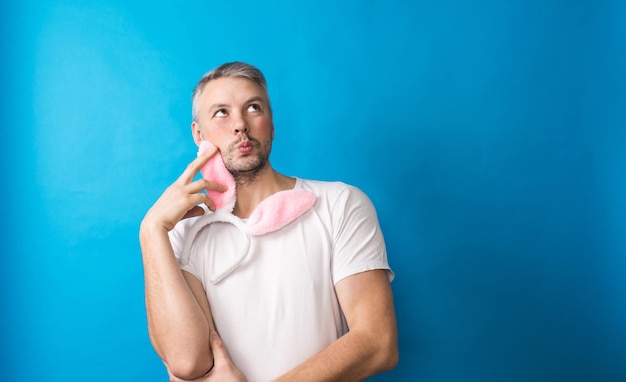 Een homoseksuele man met make-up in een wit t-shirt op een blauwe achtergrond houdt konijnenoren in zijn handen en kijkt naar de camera