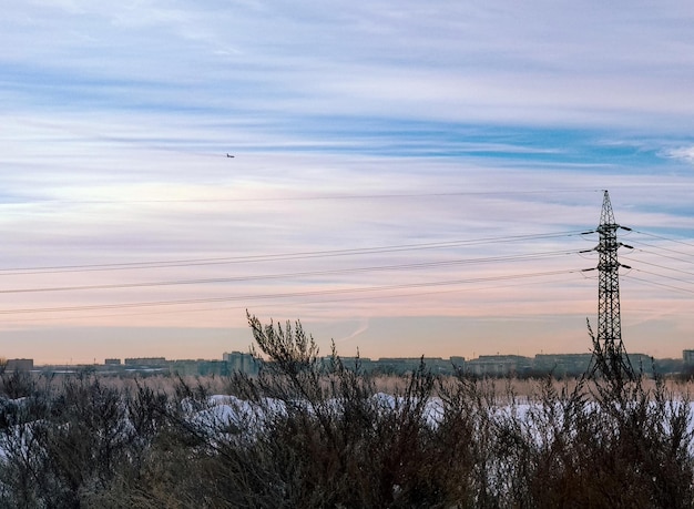 Een hoogspanningslijn staat in een besneeuwd veld tegen een panorama van de stad en een vliegtuig in de lucht