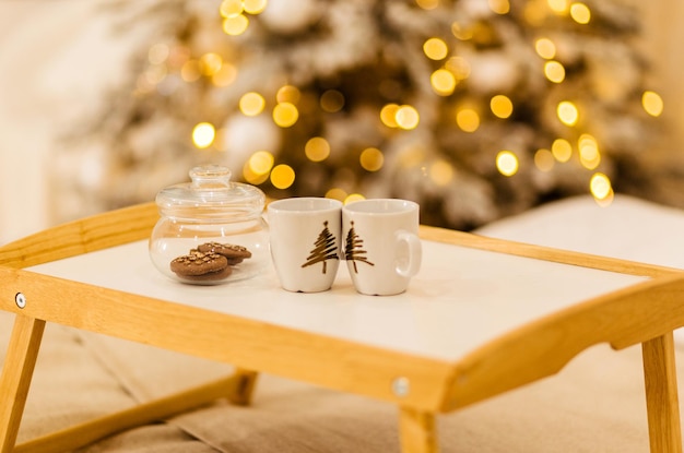 Een houten dienblad staat op het bed tegen de achtergrond van kerstverlichting. Op het dienblad staat een pot koekjes en twee kopjes