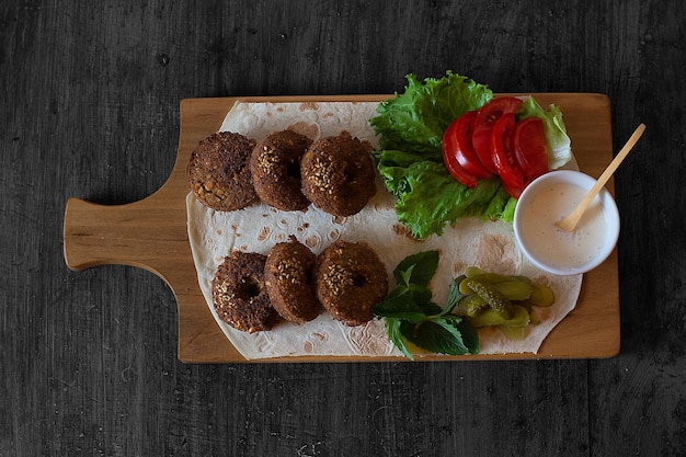 Foto een houten snijplank met een bord falafel en een salade met augurken en tomaten.