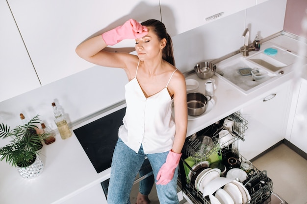 Een huisvrouw meisje in roze handschoenen na het schoonmaken van het huis zit moe in de keuken. In de witte keuken heeft het meisje de afwas gedaan en rust. Veel afgewassen vaat.