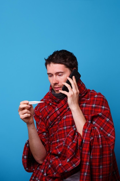 Een in een deken gewikkelde zieke man staat met een thermometer in zijn handen en roept dokter