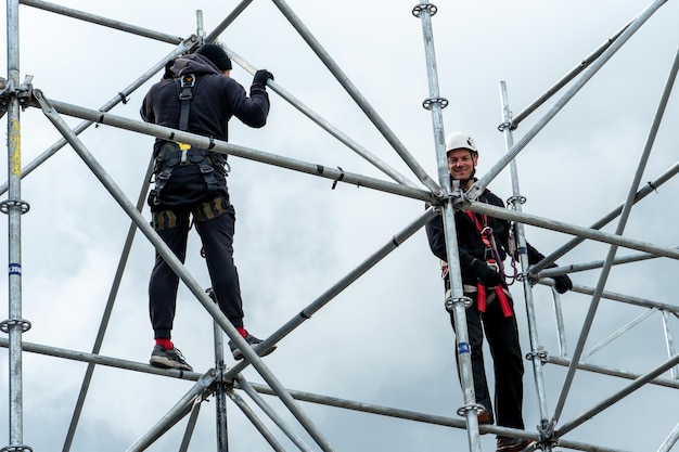 Een industriële klimmer die een helm en een beschermende riem draagt, is op grote hoogte met een touw aan een steun vastgebonden Een specialist assembleert een toneelstructuur uit een modulair systeem van metalen steigers