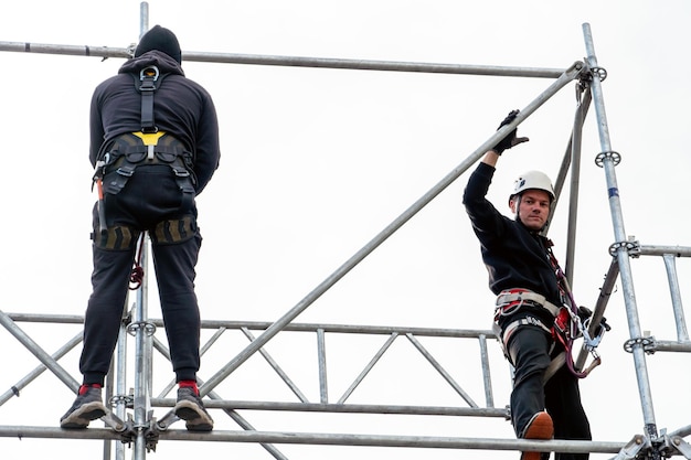 Een industriële klimmer die een helm en een beschermende riem draagt, is op grote hoogte met een touw aan een steun vastgebonden Een specialist assembleert een toneelstructuur uit een modulair systeem van metalen steigers