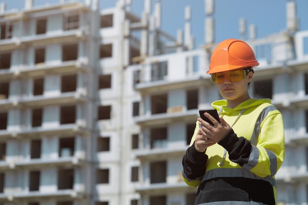 Een ingenieursmeisje in een helm en veiligheidsbril met een telefoon in haar handen tegen de achtergrond van een bouwwerf