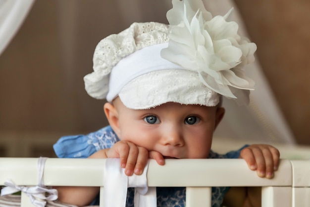 Een jaar oud kind met blauwe ogen in een stijlvolle hoed kijkt naar de camera vanuit de wieg. Kleine en schattige baby met een glimlach die in de kinderkamer zit en moeder wacht. Concept van goede opvoeding en kindertijd