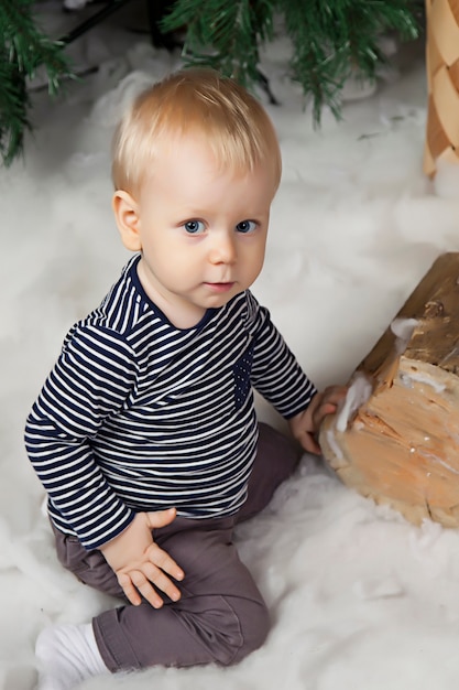Een jaar oud kind op witte deken speelt bij de kerstboom. Leuke jongen in met kerst versierde kamer