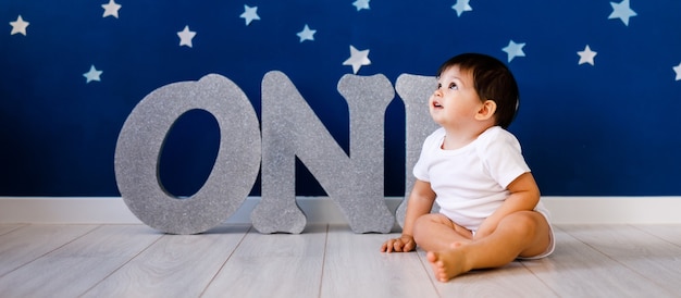 Een jaar oude babyjongen viert verjaardag in de buurt van zilveren letters ONE op blauwe achtergrond met sterren.