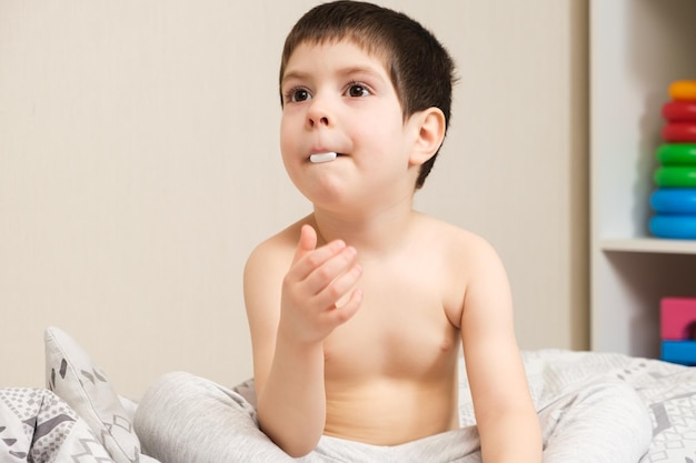 Een jarige jongen zit op een bed met een grote witte pil in zijn mond