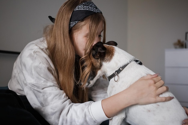 Een jong 20s meisje zit op een bed in haar woonkamer en knuffelt een kleine witte hond terwijl ze haar kust