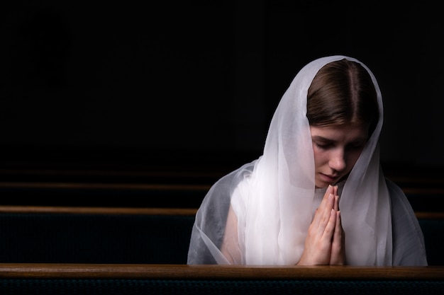 Een jong bescheiden meisje met een zakdoek op haar hoofd zit in de kerk en bidt. Het van religie, gebed, aanbidding