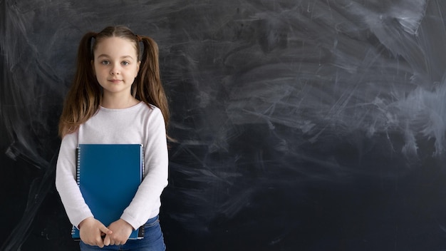 Een jong blank schoolmeisje staat tegen de achtergrond van een krijtbord en houdt verschillende notitieboekjes in zijn handen