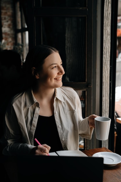 Een jong Europees freelancermeisje met lang donker haar drinkt koffie en werkt op afstand in een café