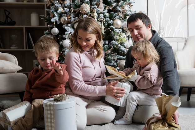 Een jong gezin met twee kleine kinderen pakt op vakantie cadeautjes uit bij de kerstboom. Vreugde en geluk in Kerstmis en Nieuwjaar.