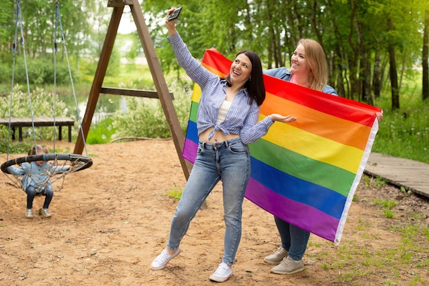 Een jong lesbisch LGBT-koppel met een kind dat lacht terwijl hij een regenboogvlag van trots vasthoudt, kijkt naar de camera