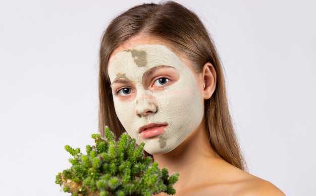 Een jong meisje, blond in een kuuroordsalon met een kleimasker op haar gezicht en een sappige cactus in haar handen. Foto op witte muur. Hoge kwaliteit foto