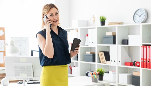 Een jong meisje dat op de telefoon in het kantoor spreekt en een glas koffie houdt.