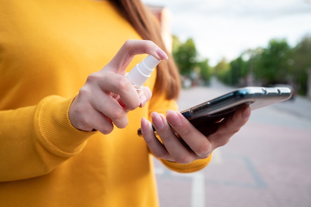 Een jong meisje in een gele trui desinfecteert haar telefoon met een reinigingsspray