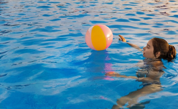 Een jong meisje in een helder luipaardzwempak, zwemt met een felgekleurde opblaasbare bal in een diepblauw zwembad met helder transparant water op een warme zonnige zomeravond