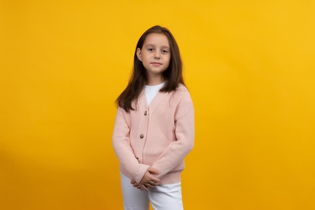 Een jong meisje in een roze jas en witte spijkerbroek poseert op een gele achtergrond