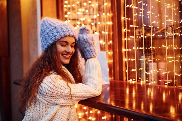 Een jong meisje in warme kleren wandelt 's avonds buiten in de stad in de buurt van een gebouw met kunstmatige feestverlichting.