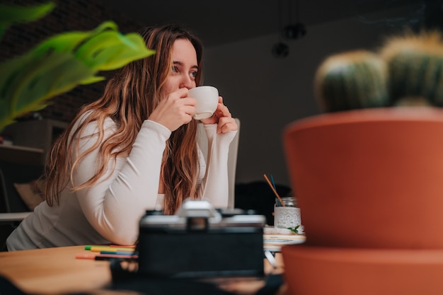 Een jong meisje koffie drinken op een houten tafel. ze heeft borstels, wierook, camera's en een groot raam.