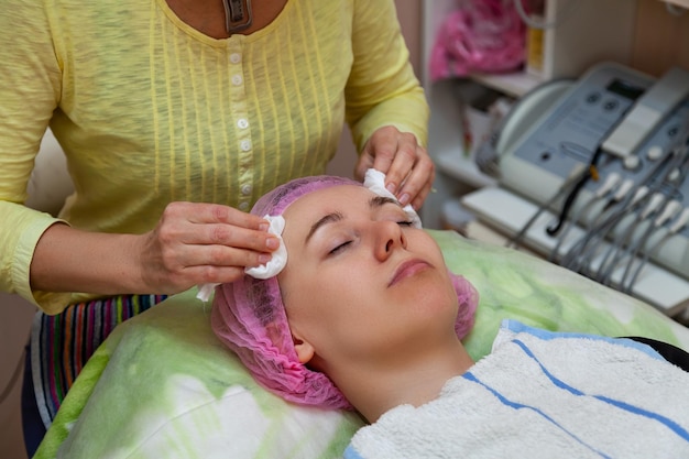 Een jong meisje ligt op een bank tijdens cosmetische ingrepen met een schoonheidsspecialiste die massage maakt Reinigende cosmetologie en make-up