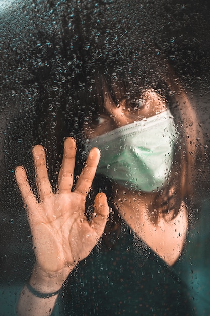 Een jong meisje met een masker in de Covid-19 pandemie kijkt uit het raam op een regenachtige dag