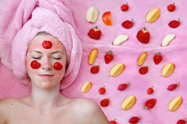 Een jong meisje met een roze handdoek ligt met haar ogen dicht met een aardbeimasker op haar gezicht. Stukjes fruit in de buurt.