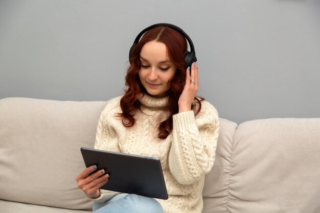 Een jong meisje met rood haar en een koptelefoon houdt een tablet in haar handen. Het meisje werkt vanuit huis