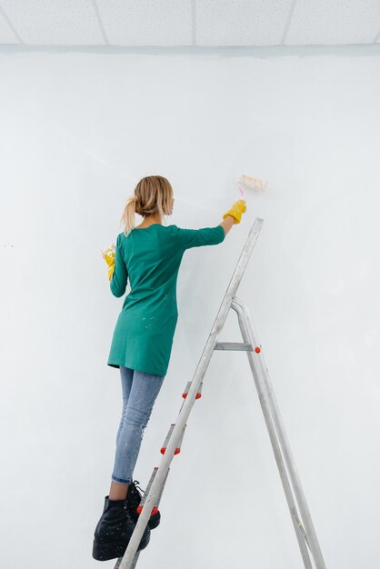 Een jong meisje op een trapladder schildert een witte muur met een roller. Reparatie van het interieur.