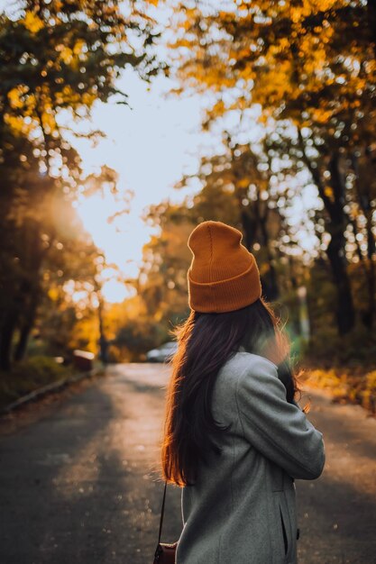 Een jong meisje staat 's avonds met zonnige stralen buiten in een kleurrijke oranje hoed alleen op straat