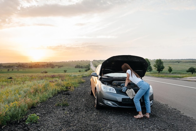 Een jong meisje staat tijdens zonsondergang in de buurt van een kapotte auto midden op de snelweg en probeert deze te repareren Het probleem oplossen Wachten op hulp Autoservice Autopech onderweg