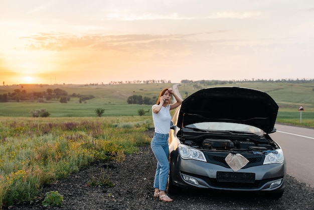 Een jong meisje staat tijdens zonsondergang in de buurt van een kapotte auto midden op de snelweg en probeert om hulp te bellen en de auto te starten Wachten op hulp Autoservice Autopech onderweg