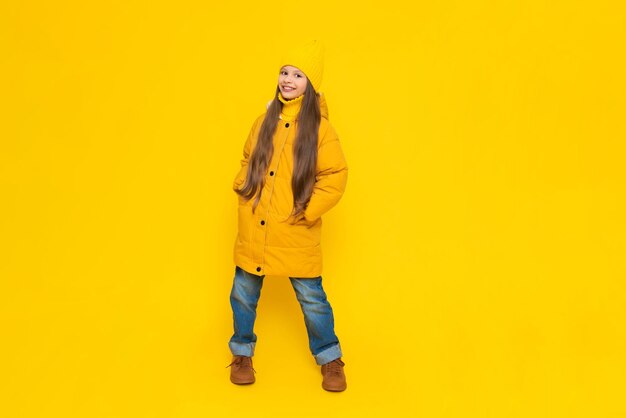 Foto een jong meisje verheugt zich bij het begin van de herfst warme kleding voor kinderen een kind in een oranje donsjack en hoed op een gele geïsoleerde achtergrond in de studio