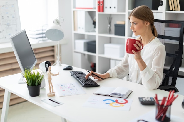 Een jong meisje zit aan een tafel op kantoor, houdt een rode kop in haar hand en werkt achter de computer.