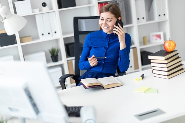 Een jong meisje zit achter een computerbureau, houdt een potlood in haar hand en praat aan de telefoon.