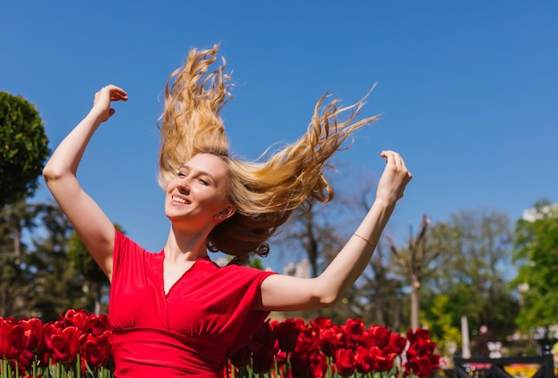 Een jong meisje zit in de zon tussen de tulpen In een rode jurk haar haren wapperen in de wind