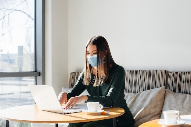 Een jong meisje zit in een café met een masker op en werkt op een computer.